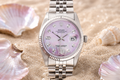 Silver watch with a pink face on sand with seashells