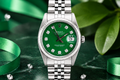 Silver watch with a green face on a dark surface with green leaves and ribbons.