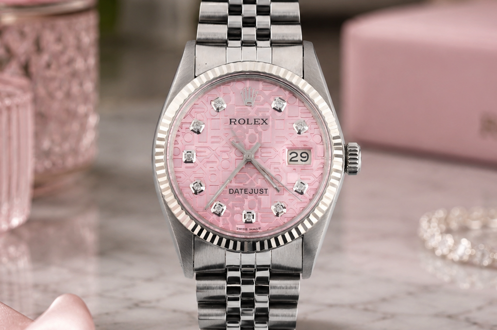 Silver watch with pink dial on a marble surface, with pink roses and jewelry box in the background.