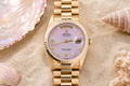 Gold watch with a pink face on sand with shells