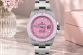 Watch with pink dial and silver case on a marble surface with flowers and box in the background